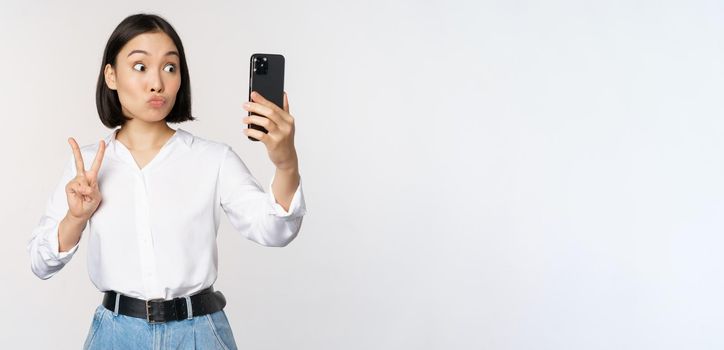 Funny asian girl showing peace, v-sign at smartphone camera, posing for photo, taking pictures with mobile application, standing over white background.