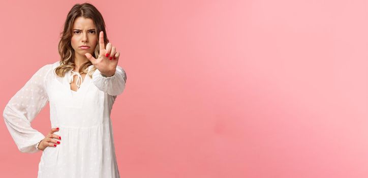 Stop, enough. Portrait of serious-looking assertive and strong blond girl in white dress, extend hand towards camera with one finger raised in warning, frowning angry, forbid and prohibit action.