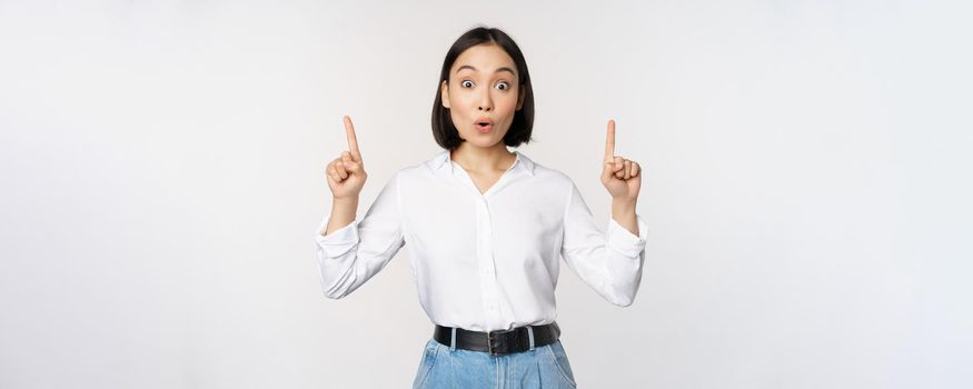 Surprised asian girl, pointing fingers up, showing top banner, information or advertisement, standing over white background in blouse. Copy space