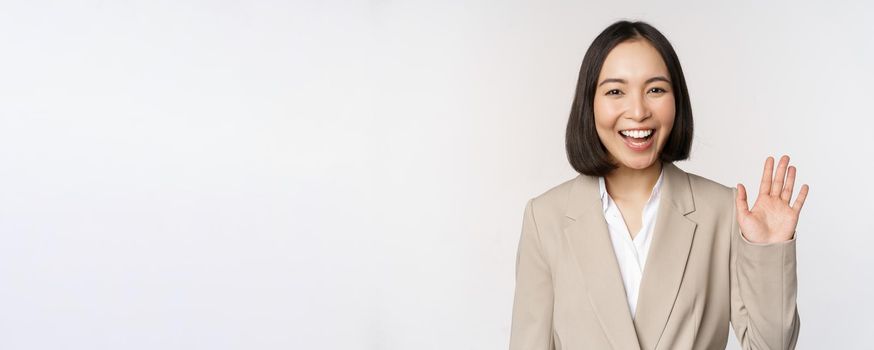 Friendly business woman, asian office lady waving hand and saying hello, hi gesture, standing over white background.