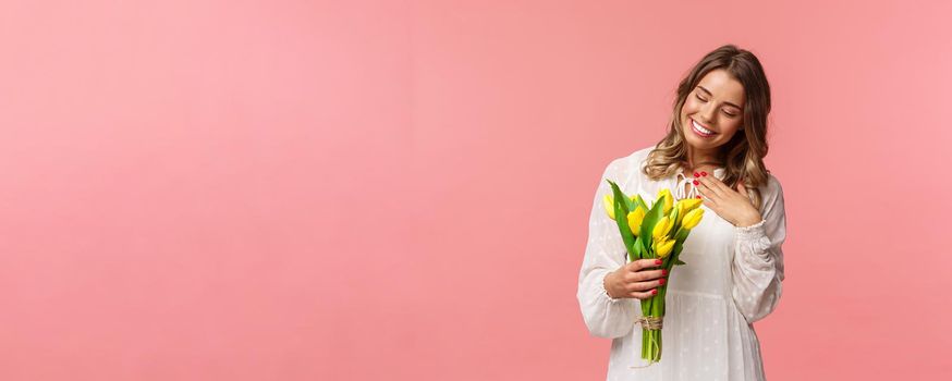 Holidays, beauty and spring concept. Portrait of pleased, cute romantic blond girl receive beautiful bouquet of flowers, looking at yellow tulips and touch heart grateful, smiling.