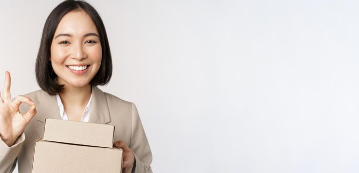 Smiling asian businesswoman, showing okay sign and boxes with delivery goods, prepare order for client, standing over white background.
