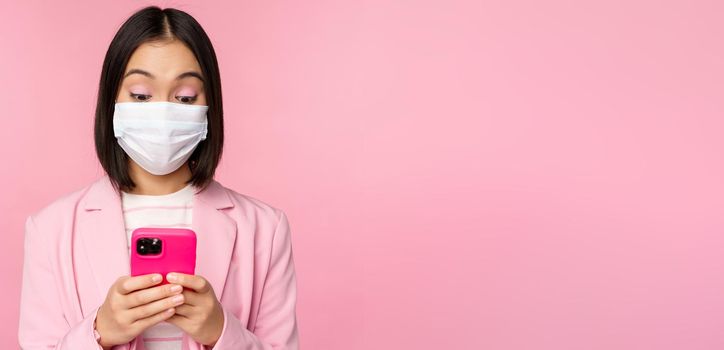 Business people and covid-19 concept. Young asian businesswoman in suit and medical face mask, using smartphone app, standing over pink background.