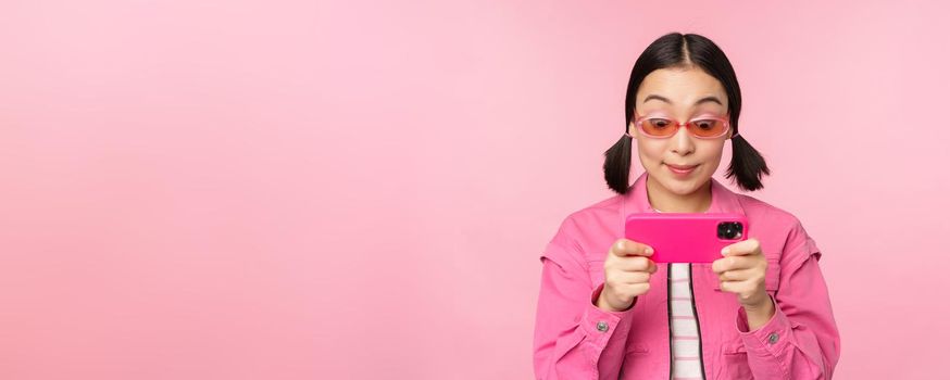 Portrait of happy asian girl playing on smartphone, watching videos on mobile phone app, standing over pink background.