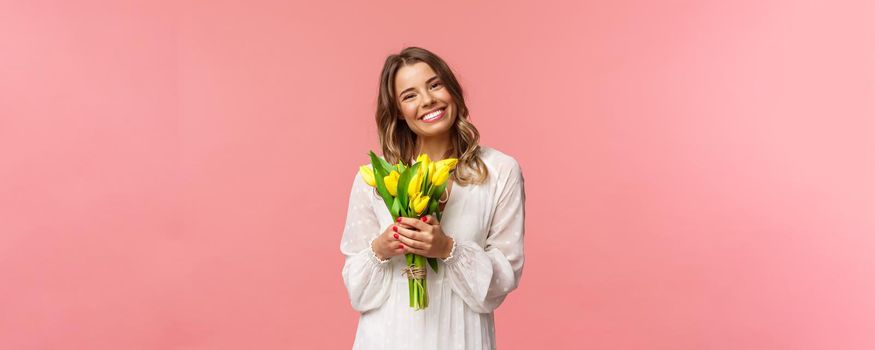 Holidays, beauty and spring concept. Portrait of lovely caucasian blond girl in white dress, smiling upbeat, holding yellow tulips, having perfect romantic date with flowers as gift, pink background.