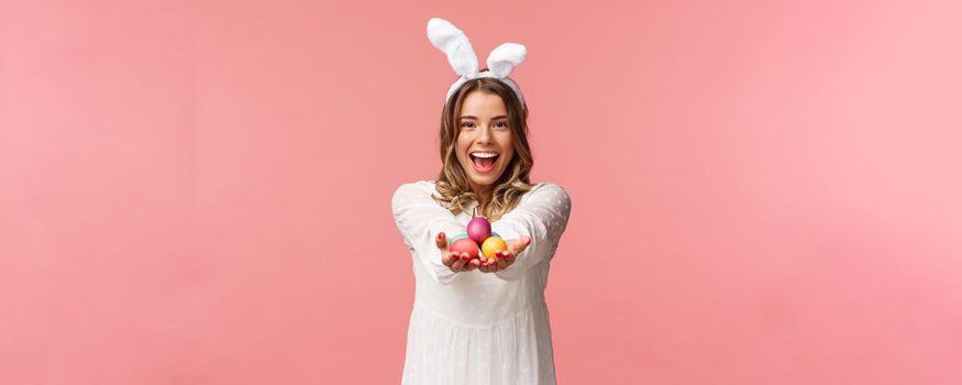 Holidays, spring and party concept. Portrait of lovely, romantic young blond woman in rabbit ears and white dress, giving you painted easter eggs as celebrating orthodox holiday, pink background.