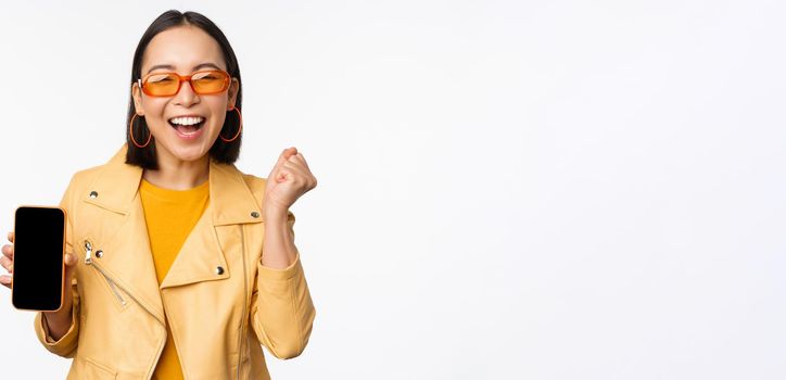 Happy asian girl in sunglasses, showing mobile phone screen, smartphone interface, laughing and smiling, celebrating, standing over white background.