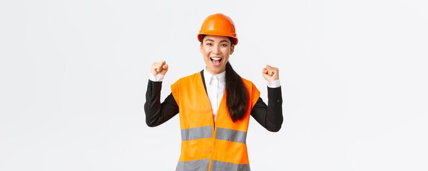 Successful winning female asian engineer fist pump and shout yes delighted, wear safety helmet and jacket, triumphing over victory, finish building in time, standing satisfied white background.