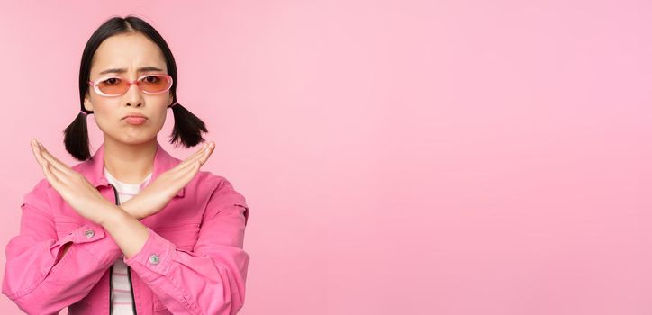 Portrait of korean girl in stylish sunglasses, sulking disappointed, showing stop, rejection gesture, cross sign, standing over pink background.