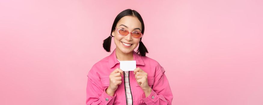 Portrait of stylish, modern asian girl, shows discount, credit card and look pleased, paying contactless, concept of shopping, standing over pink background. copy space