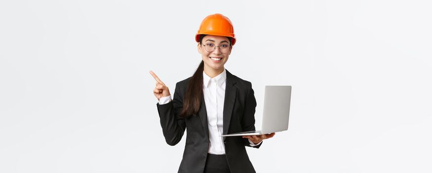 Smiling professional asian female engineer or architect at construction, wearing safety helmet and suit, pointing finger left while using laptop computer, standing white background.