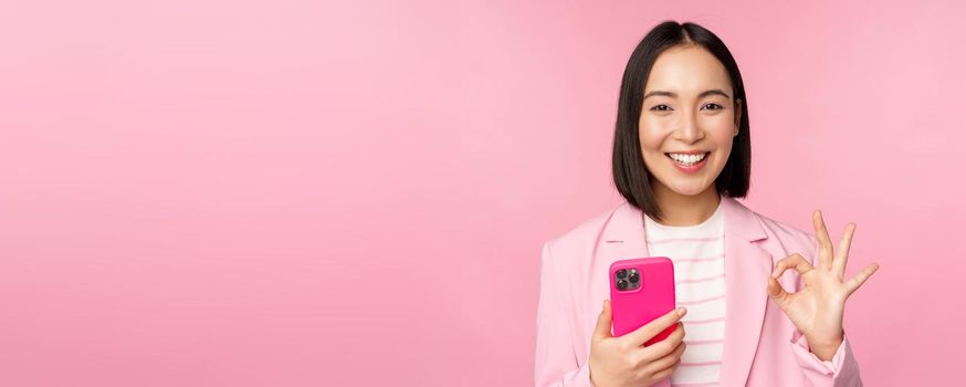 Smiling asian businesswoman showing okay sign while using mobile phone application, recommending smartphone app, standing over pink background.