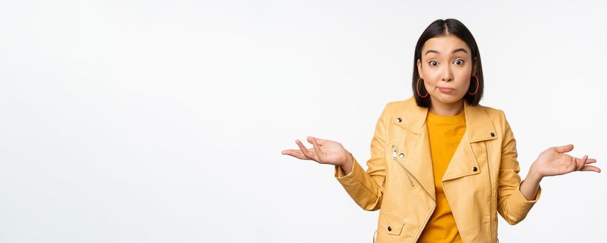 Image of korean girl shrugs shoulders and looks confused, dont know, cant tell, standing over puzzled over white background.