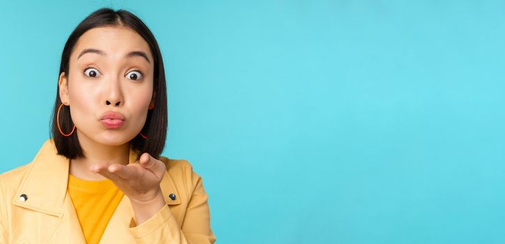 Close up portrait of funny asian girl sending air kiss, blowing at camera with popped eyes, standing over blue background.