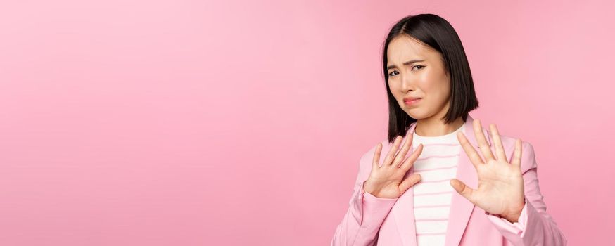 Get away. Disgusted asian businesswoman rejecting, shaking hands to refuse, grimacing from dislike, decline smth with aversion, posing in suit against pink background.