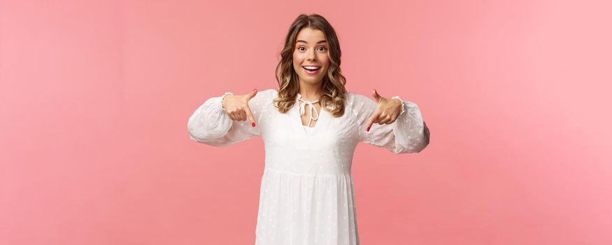 Portrait of enthusiastic upbeat young blond woman pointing fingers down to invite you check-out product, showing bottom advertisement, smiling camera cheerful, spring concept, pink background.