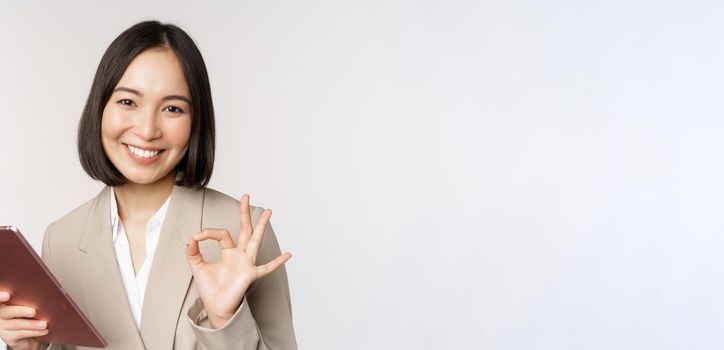 Image of successful asian businesswoman holding digital tablet, showing okay, ok sign, assuring client, standing over white background.