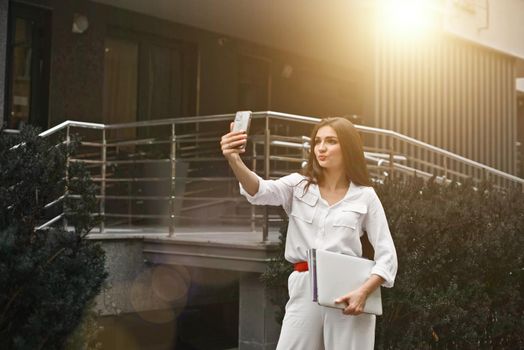 Beautiful young caucasian businesswoman in a white clothes with a laptop and notebook outdoor near business centre.