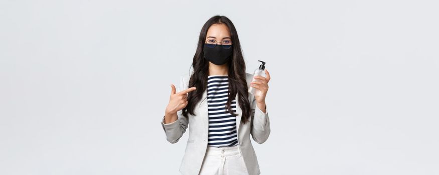 Business, finance and employment, covid-19 preventing virus and social distancing concept. Smiling cute asian office worker in face mask recommend using hand sanitizer while at work.