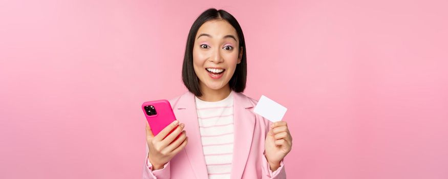 Image of smiling happy asian businesswoman showing credit card, paying online on smartphone application, order with mobile phon, standing against pink background.