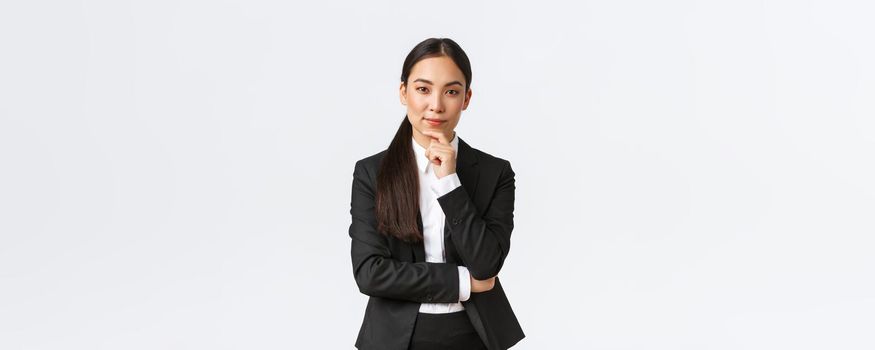 Serious pleased asian businesswoman have interesting idea, touching chin and looking cunning at camera, standing thoughtful, thinking while standing in suit over white background.