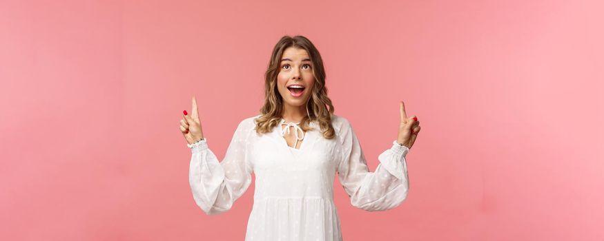 Waist-up portrait of beautiful young and healthy blond girl with cheerful, excited expression looking and pointing up, holding breath from excitement seeing something wonderful, pink background.