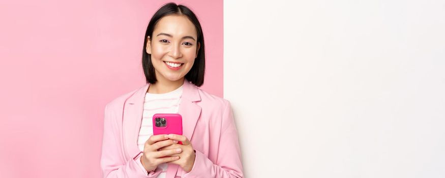 Image of korean female entrepreneur in suit, standing near info wall, advertisement on board, holding smartphone and smiling, posing over pink background.