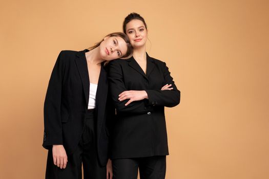 Two young business woman dressed black suit standing studio orange color background. One woman looking looking at camera arms crossed on chest other woman put her head on her shoulder. Friends support