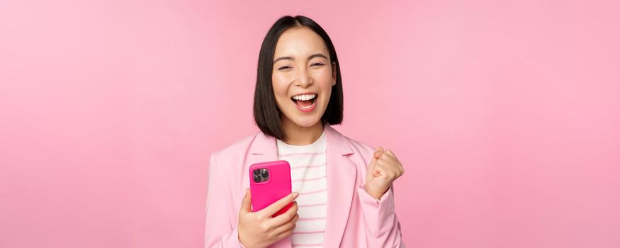 Enthusiastic asian businesswoman saying yes, winning on mobile phone, using smartphone and triumphing, celebrating success, standing over pink background.