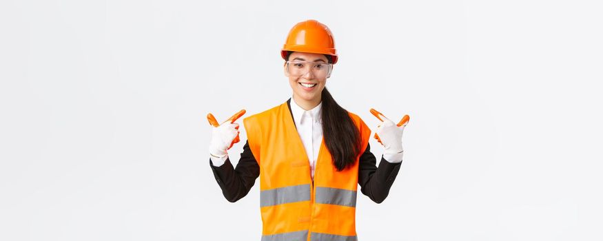 Smiling confident asian female chief construction engineer in safety helmet, gloves and glasses pointing at herself, notify to follow safe protocol and wearing special clothing, white background.