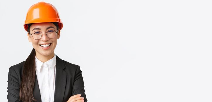 Smiling leader of the team, asian female chief engineer in safety helmet and suit cross arms confident, smiling happy at camera, introduce enterprise, standing white background.