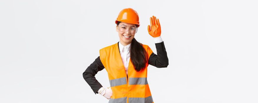 Friendly smiling asian female construction engineer, architect in safety helmet, gloves and glasses waving hand, saying hello, greeting investors or clients at building area, white background.