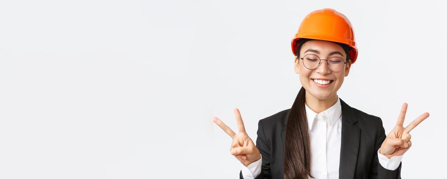 Close-up of cheerful successful female asian engineer, construction architect in safety helmet and business suit showing peace signs and smiling kawaii, standing white background.