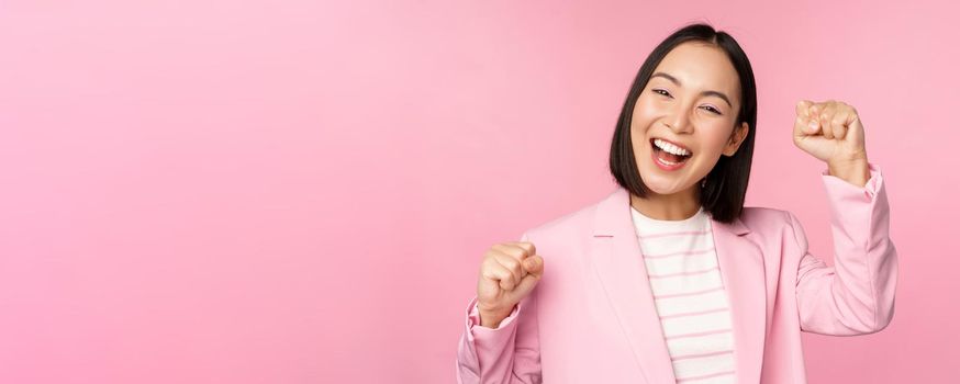 Enthusiastic asian corporate woman, businesswoman raising hand up and cheering, triumphing, winning and celebrating, standing over pink background.