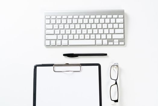 Top view of well organized workplace with office accessories. Clipboard with blank paper page, glasses and wireless keyboard on white surface. Education, creativity and working concept with copy space