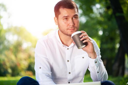 Young man sitting on green field in summer park on sunny day. Handsome man in white shirt relaxing with cup of coffee. Guy sitting on green grass and holding takeaway paper cup. Coffee break