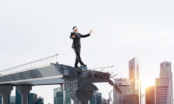 Businessman walking blindfolded on concrete bridge with huge gap as symbol of hidden threats and risks. Cityscape and sunlight on background. 3D rendering.