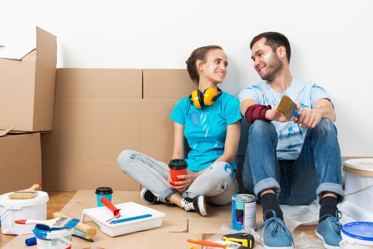 Happy smiling couple relaxing on floor with coffee after moving in new house. House remodeling and interior renovation concept. Young man and woman together sitting on floor among cardboard boxes.