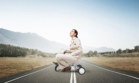 Caucasian woman riding kid's bicycle on asphalt road. Young employee in white business suit biking outdoor. metaphor of ineffective and incompetent work. Beginner level concept with bicyclist.