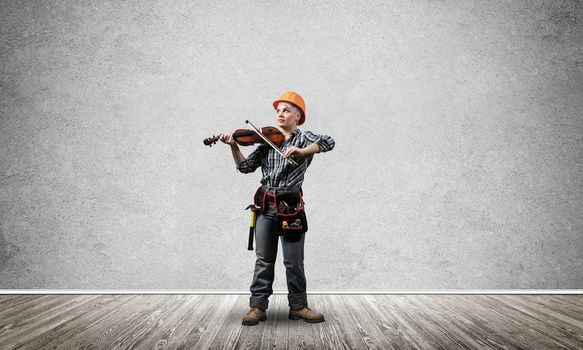 Beautiful young woman in safety helmet playing violin. Blonde female professional builder in checkered blue shirt and jeans standing in empty room. Interior design and renovation service.