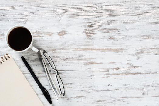 Top view office desk with white cup of coffee. Flat lay vintage wooden table with spiral notebook, pen and glasses. Coffee break time at work. Corporate business concept with copy space.