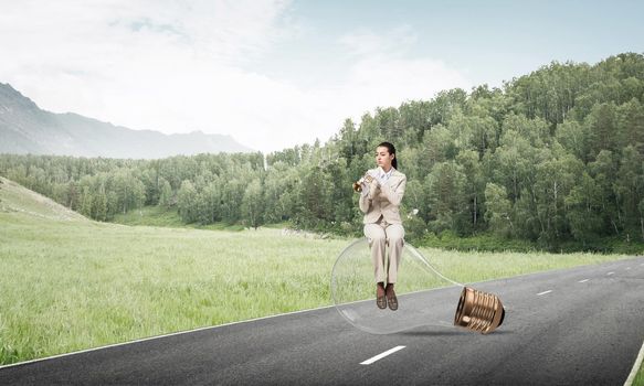 Beautiful woman playing trumpet brass on asphalt road. Young businesslady in white business suit and gloves sitting on big light bulb with music instrument. Musician practicing and performing outdoor