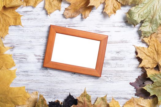 Blank rectangular photo frame lies on vintage wooden desk with bright autumn foliage. Flat lay with autumn leaves on white wooden surface. Simple brown picture frame with copy space for design.