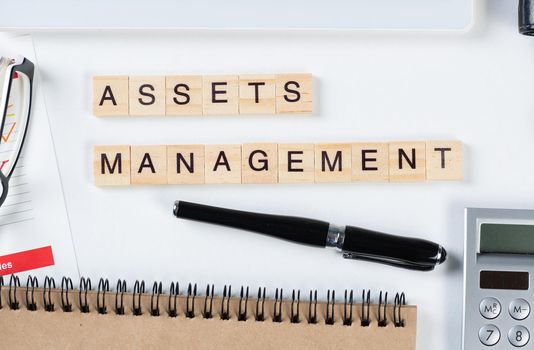 Asset management concept with letters on cubes. Still life of office workplace with supplies. Flat lay white surface with tablet computer, calculator and notepad. Investment strategy and planning.