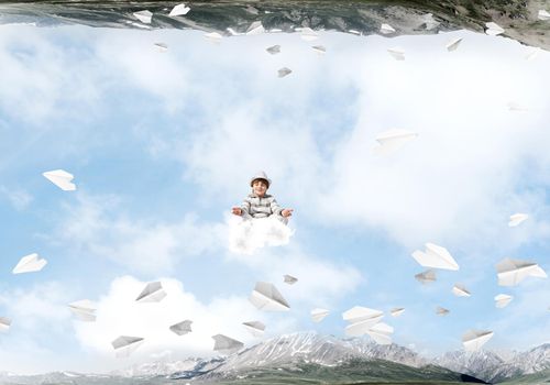 Young little boy keeping eyes closed and looking concentrated while meditating on cloud among flying paper planes and between two nature worlds.