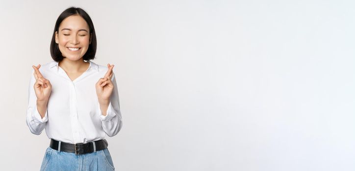 Portrait of young korean woman, asian girl cross fingers and praying, making wish, anticipating, waiting for results, standing over white background.