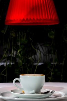 A cup of cappuccino coffee stands on the table at night under a red lampshade.