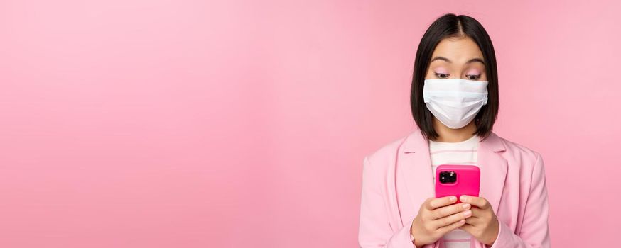 Business people and covid-19 concept. Young asian businesswoman in suit and medical face mask, using smartphone app, standing over pink background.