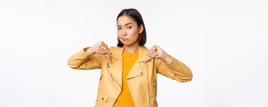 Image of fully disappointed asian woman showing thumbs down, shaking head displeased, dislike smth, standing over white background.
