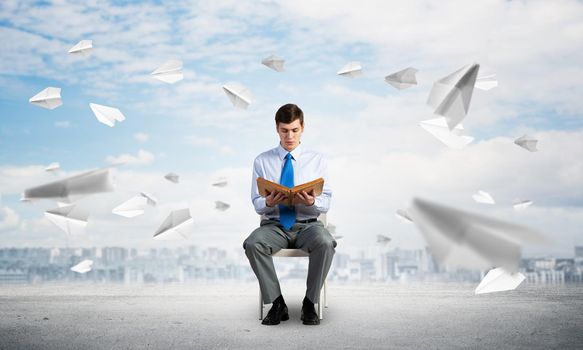 young businessman with book on office chair, paper planes fly around